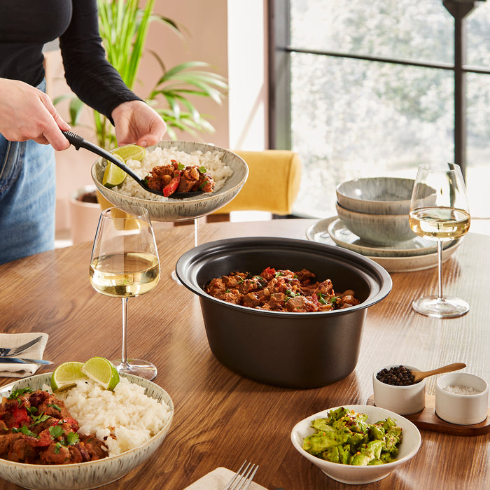 Person serving food from a slow cooker on a wooden table with wine glasses and side dishes.