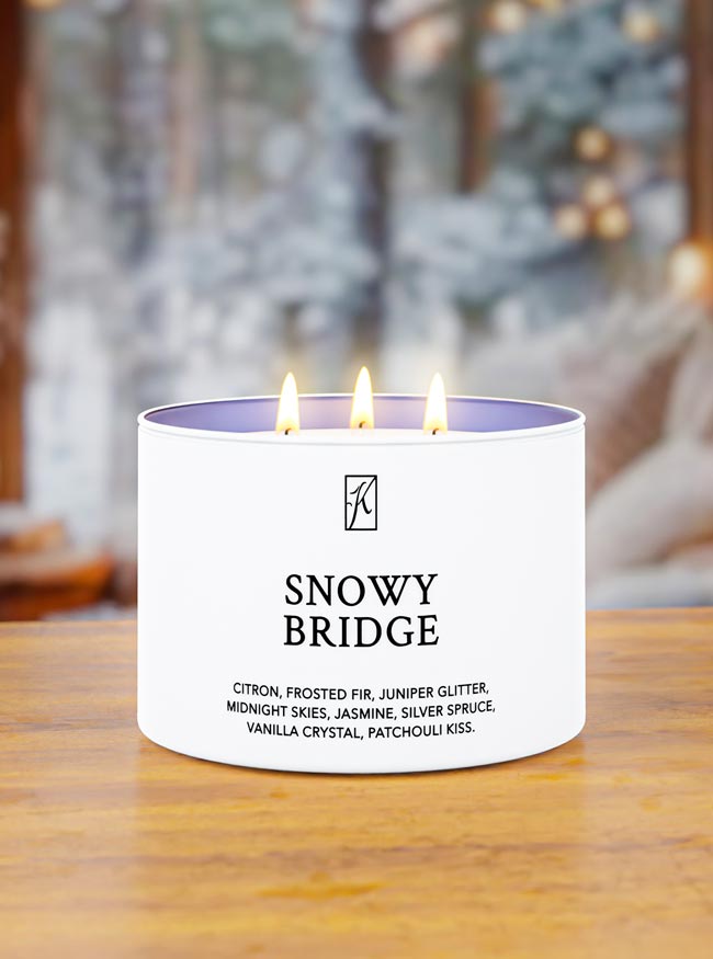 Snowy Bridge candle with two lit wicks on a wooden surface, blurred Christmas tree in the background