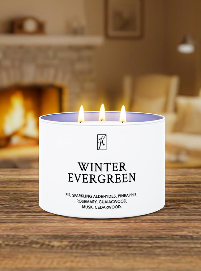 Candle labeled 'Winter Evergreen' burning on a wooden surface with a fireplace in the background