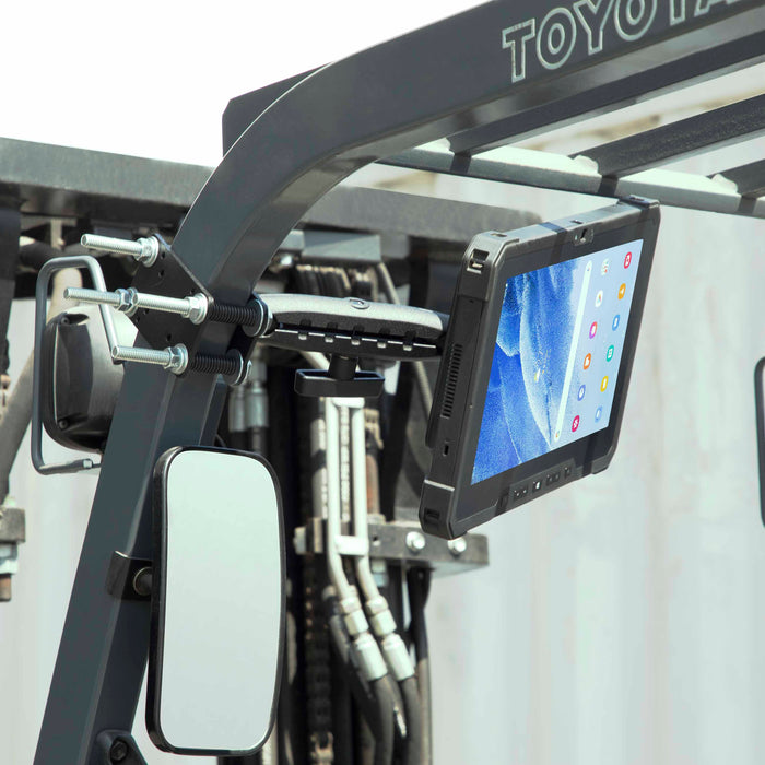 A 10.25 inch Metal Robust Forklift Front Guard VESA Mount secures a rugged tablet to the forklift’s metal frame near the rearview mirror, displaying app icons on a blue screen, with hydraulic hoses and outdoor light visible in the background.