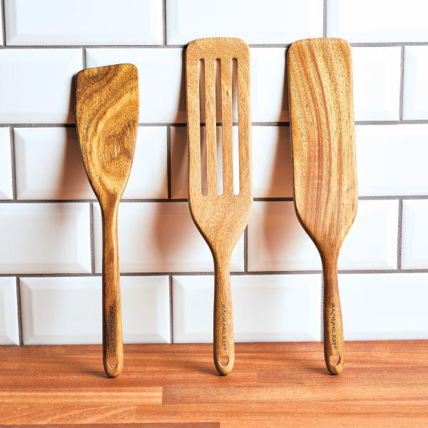 Three tools from the Acacia Wood Tool Sets are elegantly propped against a white tiled wall on a wooden countertop. These versatile spatulas, including a spurtle, are safe for use with nonstick cookware, ensuring your culinary creations remain pristine.