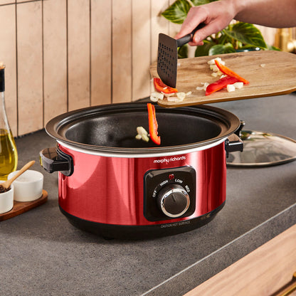 Red Morphy Richards slow cooker on a kitchen counter with a person cooking vegetables.