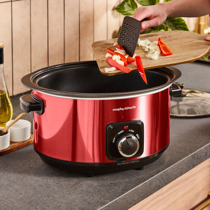 Red Morphy Richards slow cooker with food being added to it in a kitchen setting.