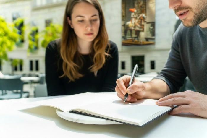 Two people sitting at a table with a notebook, one writing.