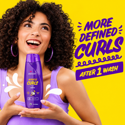 a promotional image featuring a smiling woman with defined curly hair holding aussie miracle curls shampoo in a purple bottle against a bright yellow background. bold text reads "more defined curls after 1 wash", highlighting curl definition and gentle cleansing with playful splash graphics.