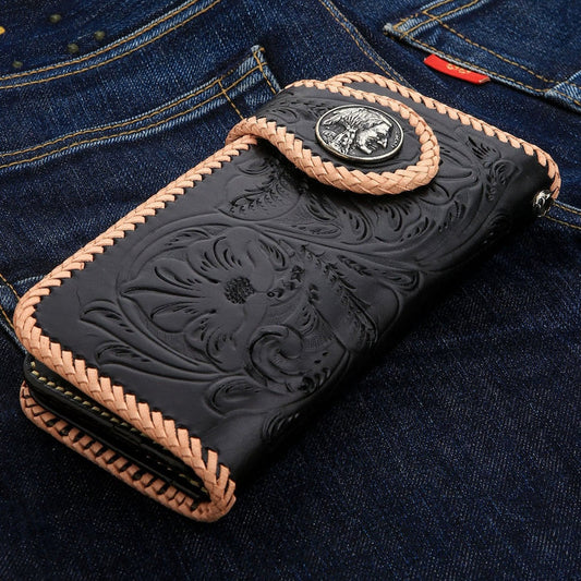 A black carved leather biker wallet with a detailed floral pattern and tan braided stitching resting on denim jeans.