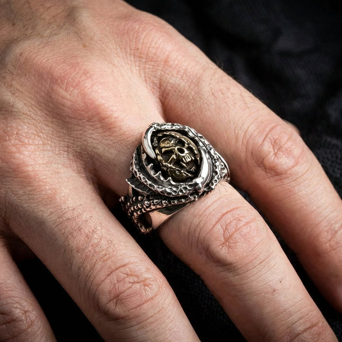 Silver claw devil ring with brass skull and crossbones centerpiece worn on man's finger, featuring detailed textured band with gothic biker style design against dark fabric background