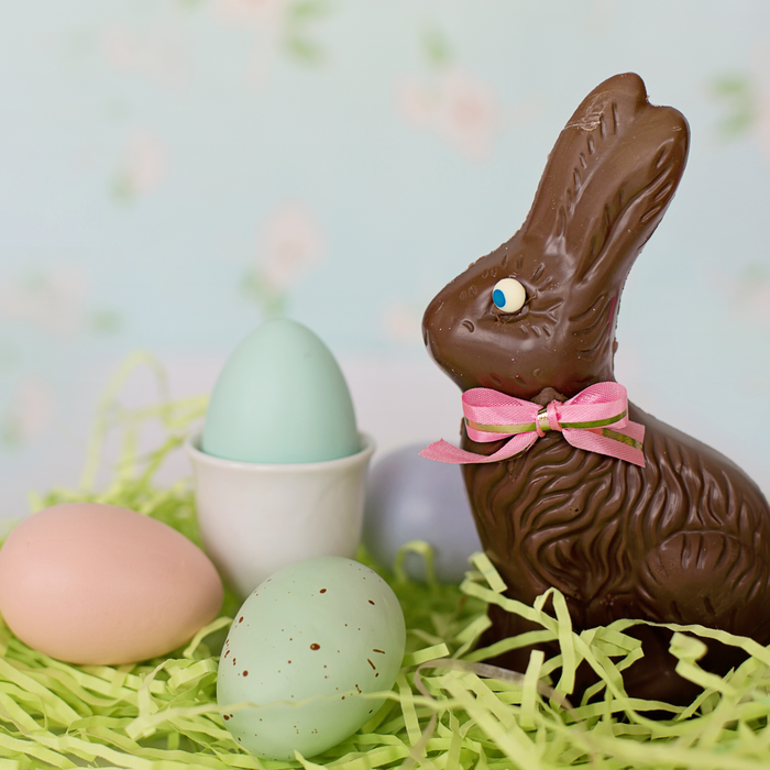 A chocolate Easter bunny with a pink and white bow sitting among colorful Easter eggs and a cup on a bed of green decorative grass.