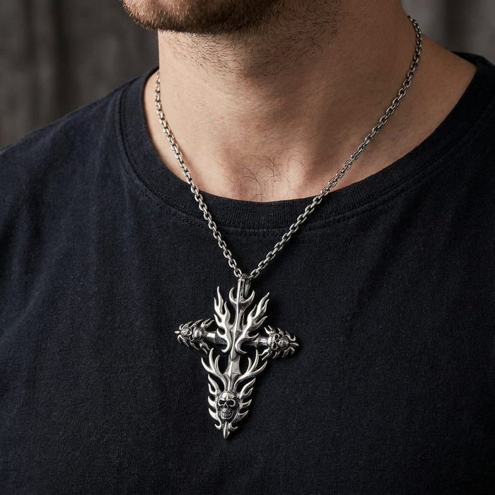 Close-up of a male model wearing a black t-shirt and a sterling silver gothic biker pendant necklace featuring a flame cross design with detailed skulls, hanging on a silver chain.