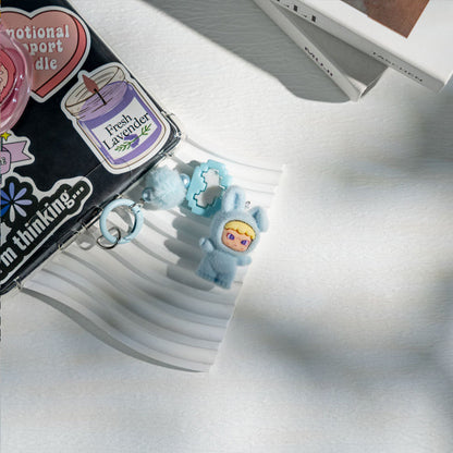 Small blue character charm on a white surface with a black notebook and stickers in the background.