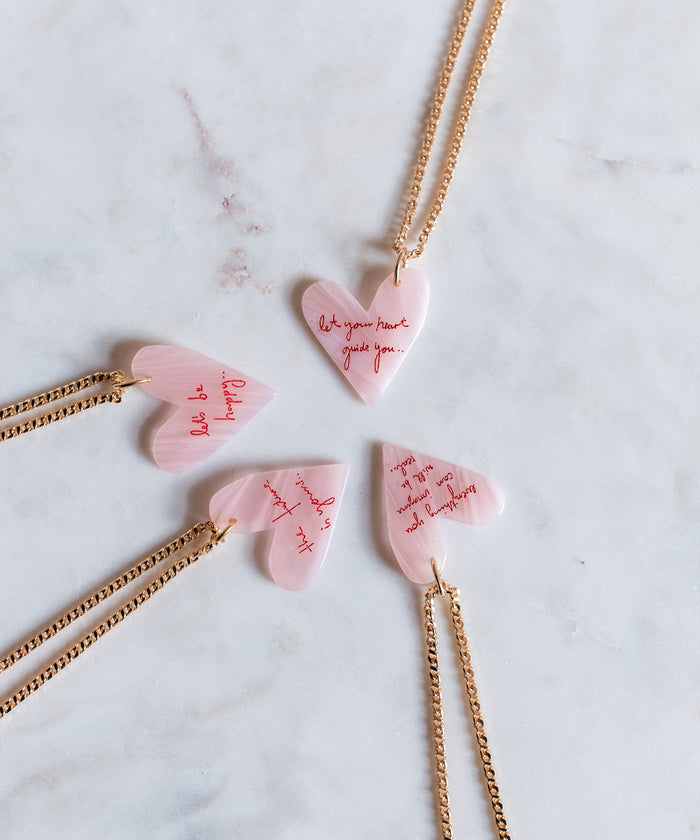 Four "Let's be happy" gold necklaces by WALD Berlin form a circle. Each 18k gold-plated, pink heart-shaped pendant is hand-painted with a unique red inscription that exudes positivity. They are elegantly arranged on a light marble surface.
