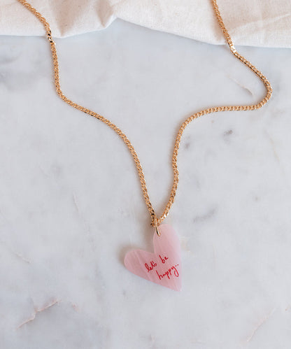 The WALD Berlin "Let's be happy" gold necklace features an 18k gold-plated chain with a pink heart-shaped hand-painted pendant inscribed with "don't be happy," stylishly displayed on a marble surface.
