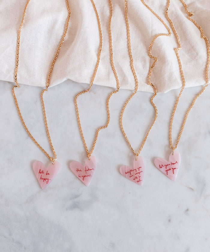 The WALD Berlin Let's be happy gold necklaces feature pink heart pendants with motivational messages, elegantly resting on a marble surface against a white polyester backdrop. Made in Germany, each piece radiates unique charm and craftsmanship.