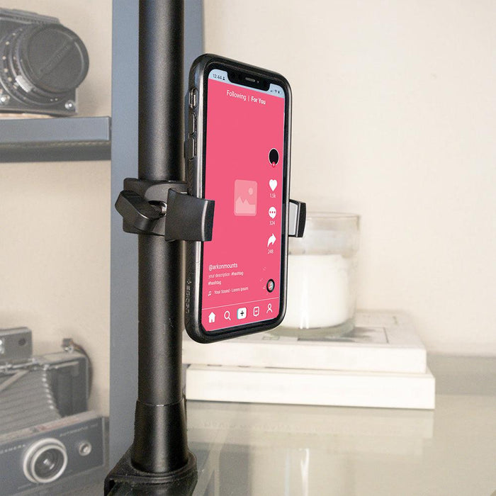 A smartphone is mounted vertically in the Mobile Grip 5 Phone Holder with Clamp Mount, attached to a pole. The screen shows a social media app with a pink background; cameras, a white candle, and stacked books are visible behind it.