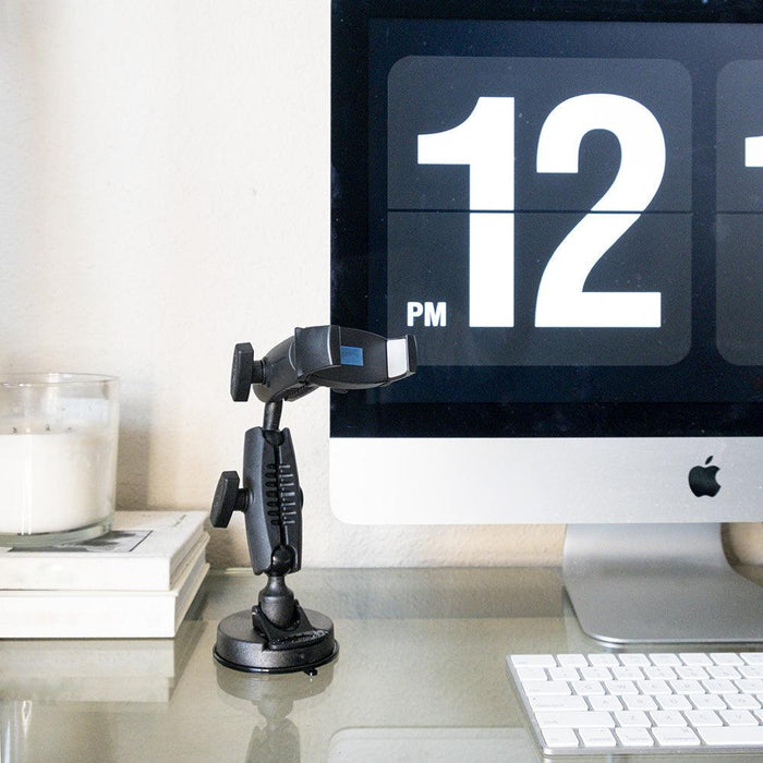 The Mobile Grip 5 Robust Phone Mount Holder for iPhone, Galaxy, and Note sits on a desk beside a candle, stacked books, and an iMac showing 12 PM. A wireless keyboard is on the glass surface. Ideal for hands-free use or close-up video recording.