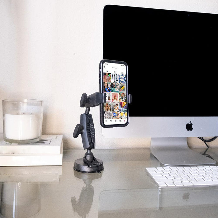 The Mobile Grip 5 Robust Phone Mount Holder securely displays a smartphone with photos on a glass desk next to an Apple iMac, wireless keyboard, candle, and books in a modern workspace.