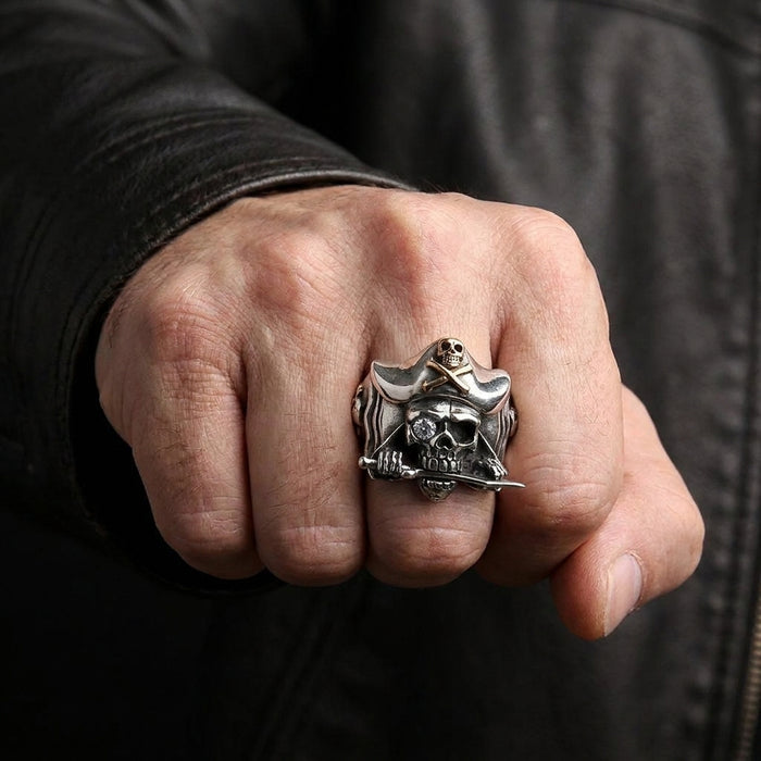 Close-up lifestyle shot of a man's fist wearing a chunky silver pirate skull ring featuring a tricorn hat, a clear gemstone eye, and a sword in its teeth, styled against a black leather jacket.