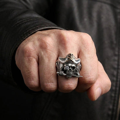 Close-up lifestyle shot of a man's fist wearing a chunky silver pirate skull ring featuring a tricorn hat, a clear gemstone eye, and a sword in its teeth, styled against a black leather jacket.