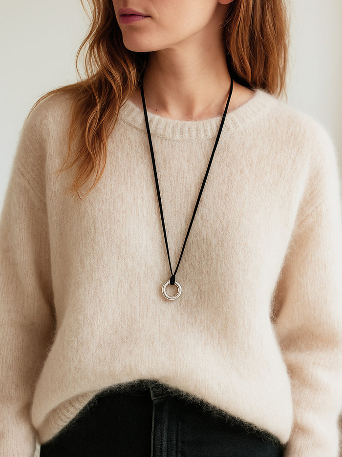 Kvinna som bär ett långt halsband med silverring i läderband utanpå en vit stickad oversized tröja.
