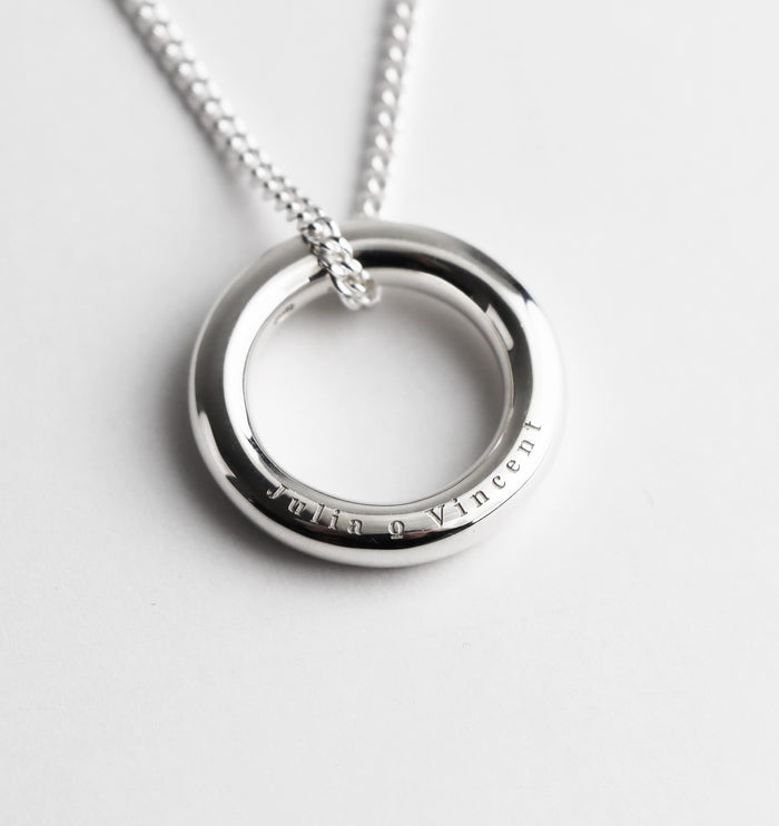 Silverring med två namn graverade på sidan av ringen. Ringen hänger i en halskedja, pansarkedja.