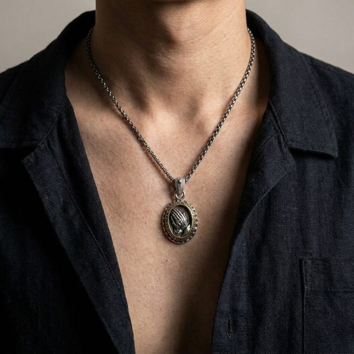 Close-up lifestyle shot of a man wearing a vintage-style silver oval praying hands pendant on a silver chain necklace, styled with an open dark linen shirt against a neutral background.
