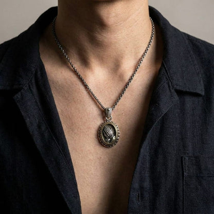 Close-up lifestyle shot of a man wearing a vintage-style silver oval praying hands pendant on a silver chain necklace, styled with an open dark linen shirt against a neutral background.