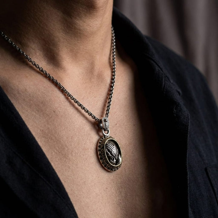 Close-up of a male model wearing an oxidized silver oval praying hands pendant on a sturdy chain necklace, styled with an unbuttoned black shirt.