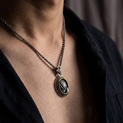 Close-up of a male model wearing an oxidized silver oval praying hands pendant on a sturdy chain necklace, styled with an unbuttoned black shirt.
