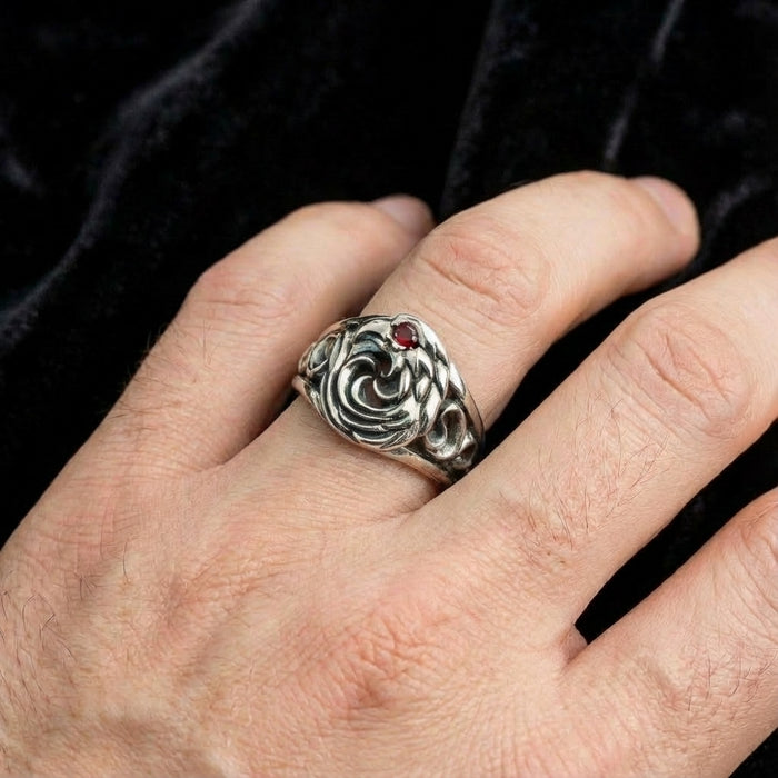 Close-up of a men's sterling silver eagle ring featuring a red gemstone eye, worn on the ring finger against a dark velvet background.