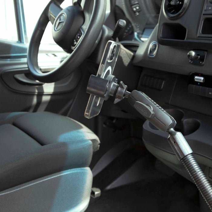 A smartphone is mounted on the RoadVise® Heavy-Duty Seat Rail or Floor Phone Mount near the driver’s seat and steering wheel, with the car’s interior and dashboard controls visible in the background.
