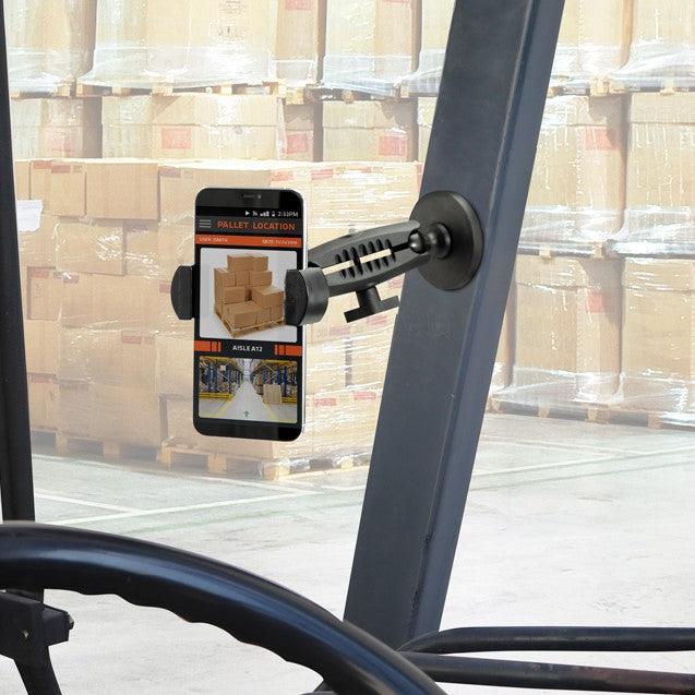 A smartphone is secured with the RoadVise® Holder with Magnetic Mount inside a forklift, displaying a pallet location app and a photo of stacked boxes while large cardboard box stacks fill the background warehouse.