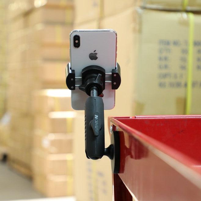 A RoadVise® Holder with Magnetic Mount secures a smartphone with handle to the side of a red metal surface, while stacks of cardboard boxes appear blurred in the background.