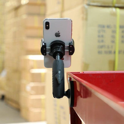 A RoadVise® Holder with Magnetic Mount secures a smartphone with handle to the side of a red metal surface, while stacks of cardboard boxes appear blurred in the background.
