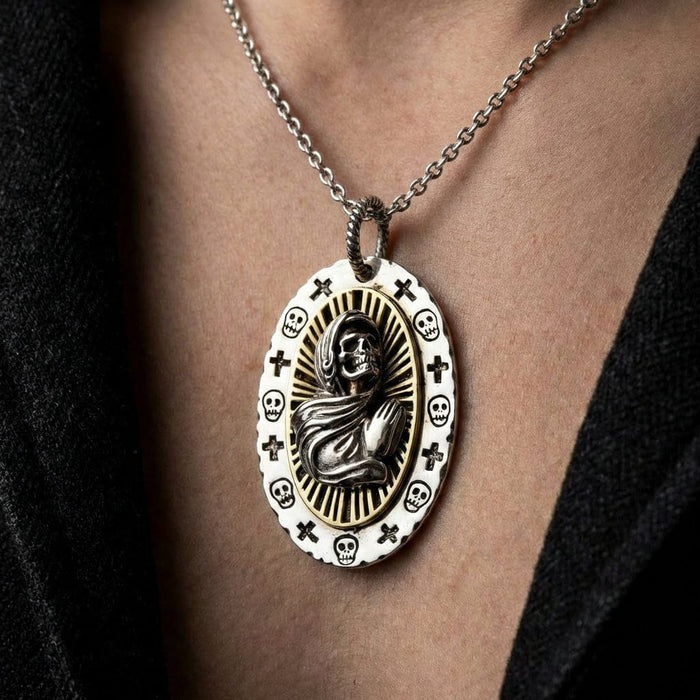 Santa Muerte silver pendant necklace featuring a robed skeleton figure with radiating halo design, surrounded by skulls and crosses on white enamel border, worn on silver chain against skin with black clothing