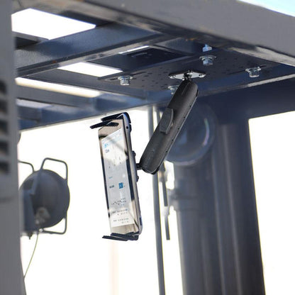 A Slim-Grip® Tablet Holder with Forklift Overhead Guard Mount and 6 Metal Shaft secures a smartphone upside down on a black metal bracket attached to a steel frame, with blurred industrial equipment in the background.
