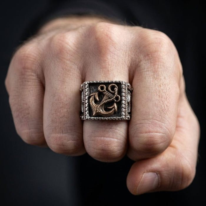 Men's anchor Viking signet ring with bronze anchor motif on oxidized silver square face, worn on middle finger of a clenched fist against dark background