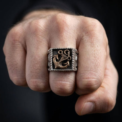 Men's anchor Viking signet ring with bronze anchor motif on oxidized silver square face, worn on middle finger of a clenched fist against dark background