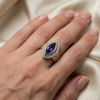 Close-up of a woman's hand wearing a vintage-style gold bishop ring featuring a large marquise-cut amethyst center stone surrounded by a halo of small light blue gems against a soft beige satin background.